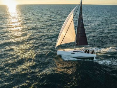 Segelboot mit dunklem Großsegel, drei Personen an Bord, Sonnenuntergang über dem Meer