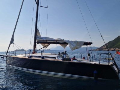 Segelboot im Meer, Mast und aufgewickelte Segel; Küstenlandschaft im Hintergrund.