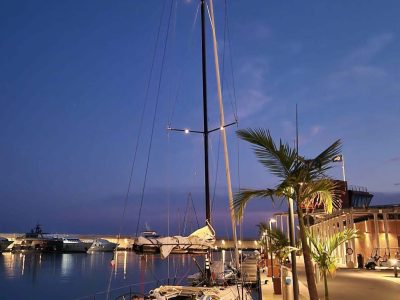 Segelboot am Kai im Hafen bei Dämmerung, Palmen und Wasserreflexionen.
