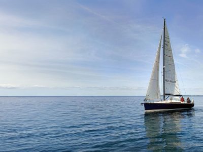 Segelboot mit weißen Segeln auf ruhigem blauem Meer, zwei Passagiere an Bord.